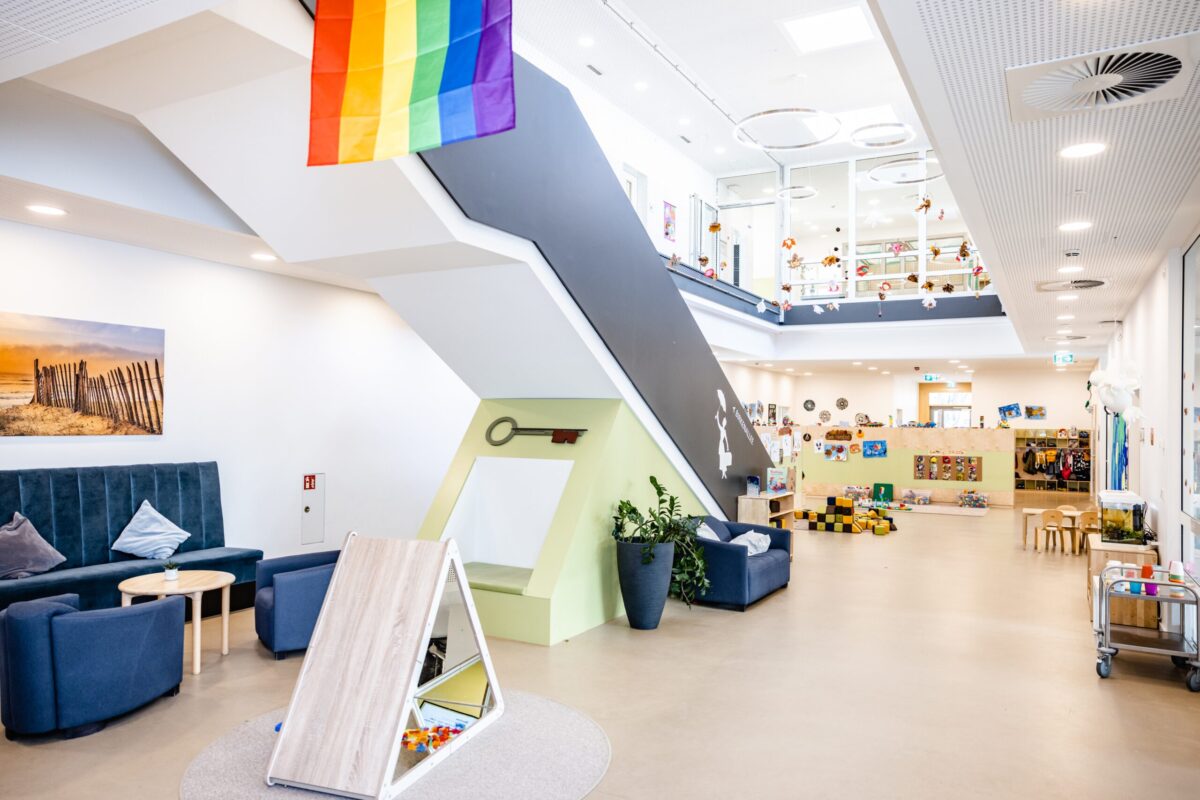 Bunt dekorierte Kita mit Spielbereichen und einem großen Treppenaufgang; eine Regenbogenflagge hängt von der Decke.