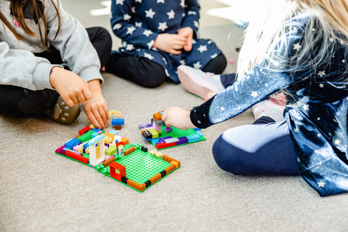 Kinder spielen mit bunten Bauklötzen auf dem Teppich; eine Hand fügt Teile hinzu, während andere Hände ebenfalls am Spiel beteiligt sind.