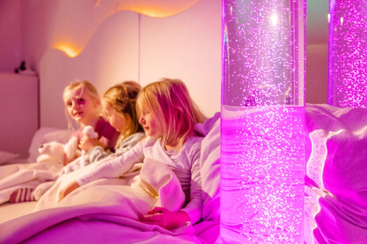 Drei Mädchen sitzen in einem Bett, umgeben von Decken; sie kuscheln mit Plüschtieren und schauen auf einen beleuchteten Wasserzylinder, der pinke Blasen zeigt.