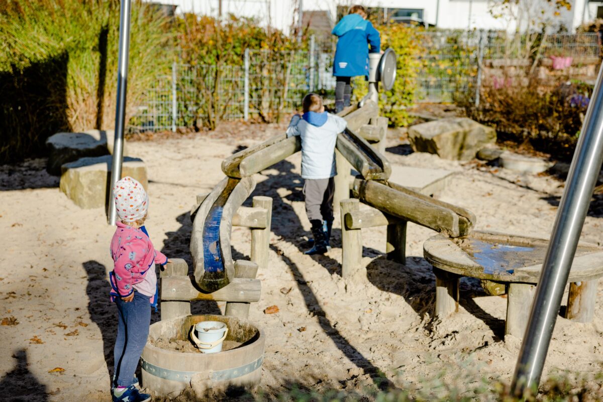 Zwei Kinder spielen in einem Sandspielbereich mit einer Holzspielkombination; ein Kind erkundet eine Wasserbahn, während das andere auf einem Kletterelement steht.